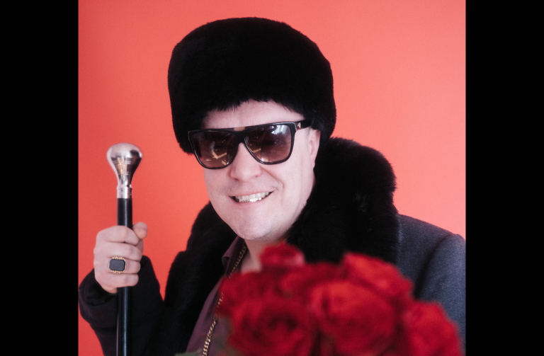 Der Rapper Morlockk Dilemma mit Sonnenbrille, einen Strauß Rosen, edlen Gehstock und in schwarzer Mütze, Mantel und Sonnenbrille.