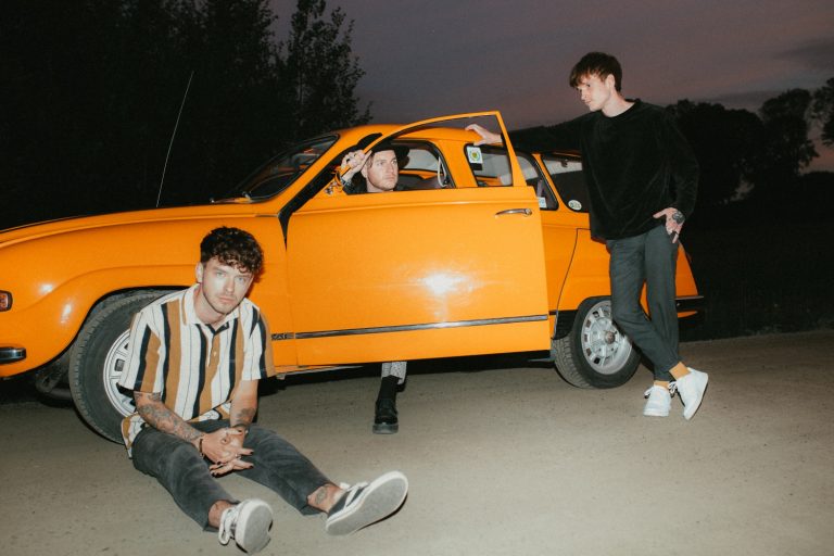Mitglieder der Band Normandie sitzen/stehen um/in orangefarbenen Auto mit geöffneter Fahrertür