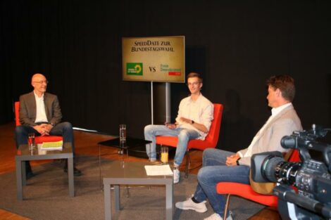 Andreas Tesche, Moderator Konrad und Hagen Reinhold sitzen im Halbkreis Richtung Kameras. Im Hintergrund steht ein Monitor mit der Aufschrift "SpeedDate zur Wahl: GRÜNE vs FDP"