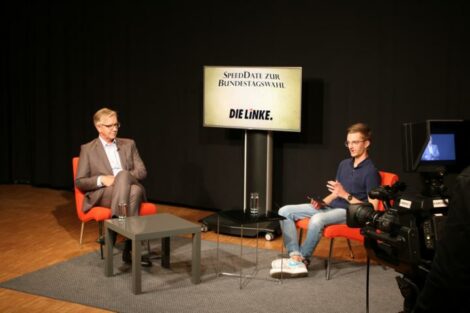 Dietmar Bartsch und Konrad sitzen sich schräg gegenüber. Hinter ihnen ein Monitor mit der Aufschrift "SpeedDate zur Wahl: Die Linke".