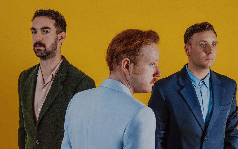 Mitglieder der Musikband Two Door Cinema Club (Kevin Baird, Alex Trimble, Sam Halliday) vor gelben Hintergrund