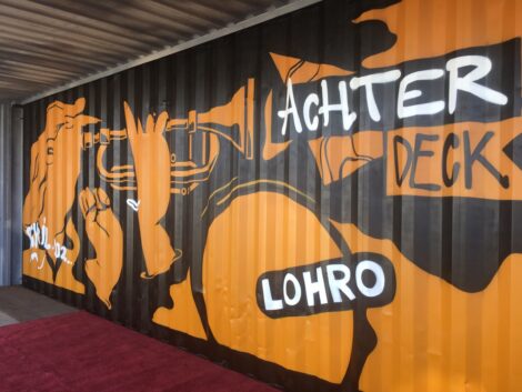 Schiffscontainer mit schwarz-orangenem Graffiti: Trompete-spielende Person mit Schriftzug "Achterdeck" davor und Logo von Radio LOHRO darunter