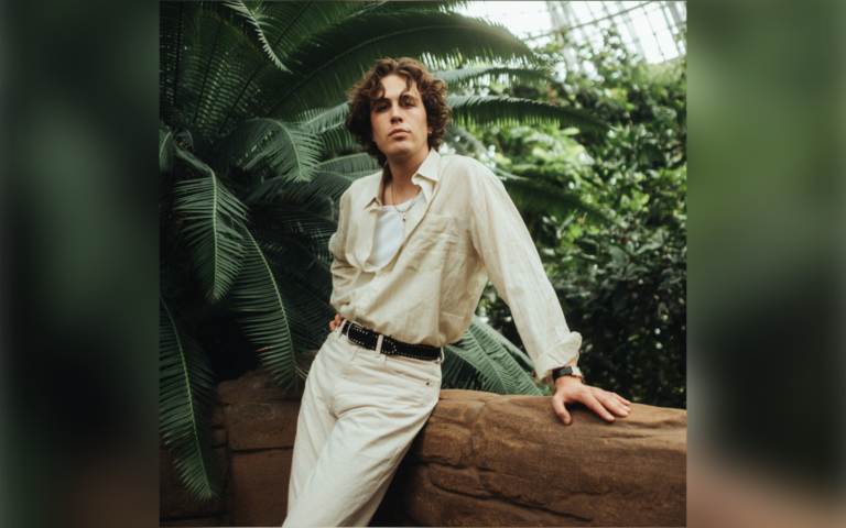 Künstler Fil Bo Riva in naturfarbiger Hose und Hemd lehnt an Mauer vor tropischen Pflanzen in einer Halle mit Glasdach