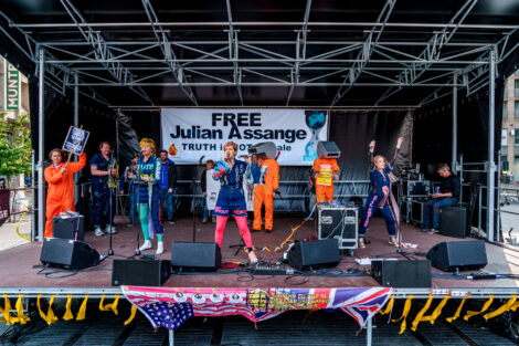 Frontansicht einer Bühne mit mehreren Künstlern, Banner im Hintergrund "Free Assange – Truth is NOT for sale"