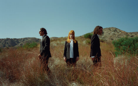 Drei Personen der Band Paramore nebeneinander stehend in schwarzen Anzügen und weißen Hemden in Gras-/Busch-Landschaft bei sonnigem Wetter und blauem Himmel. Linke Person in nach links schauend, rechte ebenso nach rechts, dazwischen Sängerin Hayley Nichole Williams mit Blick in die Kamera.