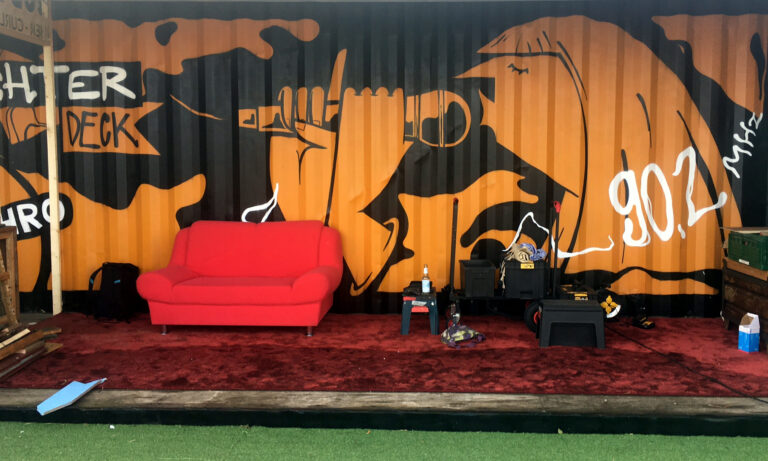 Seitenansicht von Schiffscontainer, der mit schwarz-orangenem Graffiti gesprayt ist, wo eine Frau in Seitenansicht ein Mikrofon mit beiden Händen vor dem Mund hält. Davor steht ein rotes Sofa und andere Dekoration