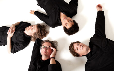 Vier Mitglieder der Musikgruppe STRFKR liegen in schwarzer Kleidung auf dem Rücken auf weißem Boden; Köpfe beieinander, Körper in alle vier Himmelsrichtungen abgehend