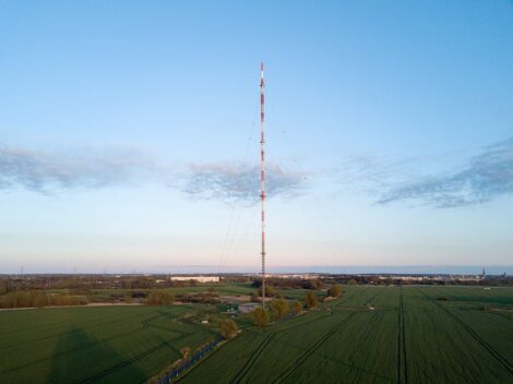 Rot-weiß gefärbter Stahlgitter-Sendemast vor klarem Himmel auf grünem Feld