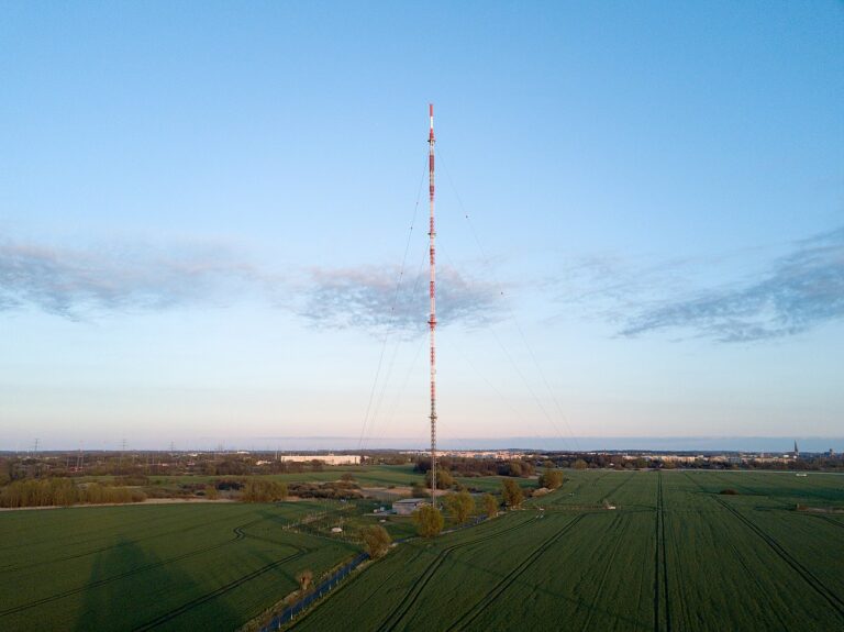 Rot-weiß gefärbter Stahlgitter-Sendemast vor klarem Himmel auf grünem Feld