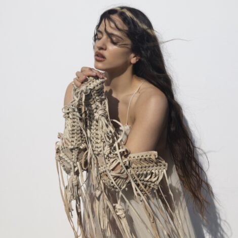 Die Künstlerin ROSALÍA steht vor einem hellen Hintergrund. Sie trägt ein beiges Kleid mit Schnüren und Fransen an den Armen.
