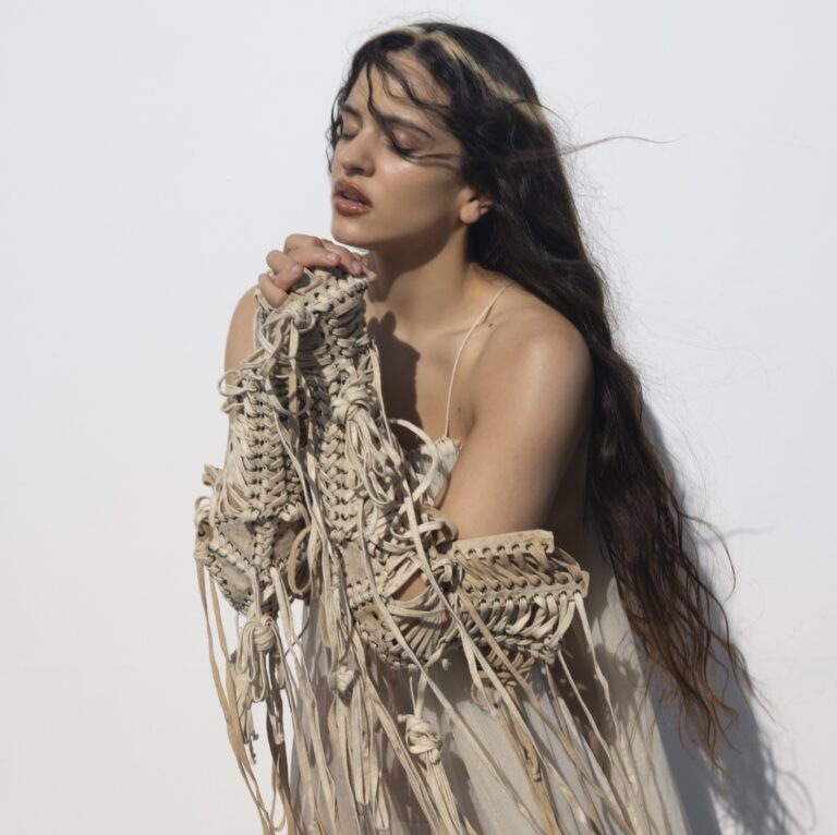 Die Künstlerin ROSALÍA steht vor einem hellen Hintergrund. Sie trägt ein beiges Kleid mit Schnüren und Fransen an den Armen.