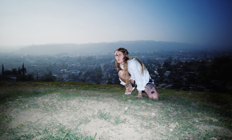 Musikerin Lala Lala kniet auf einem mit Gras bewachsenen Hügel, im Hintergurnd ist die Silhouette einer Stadt zu sehen